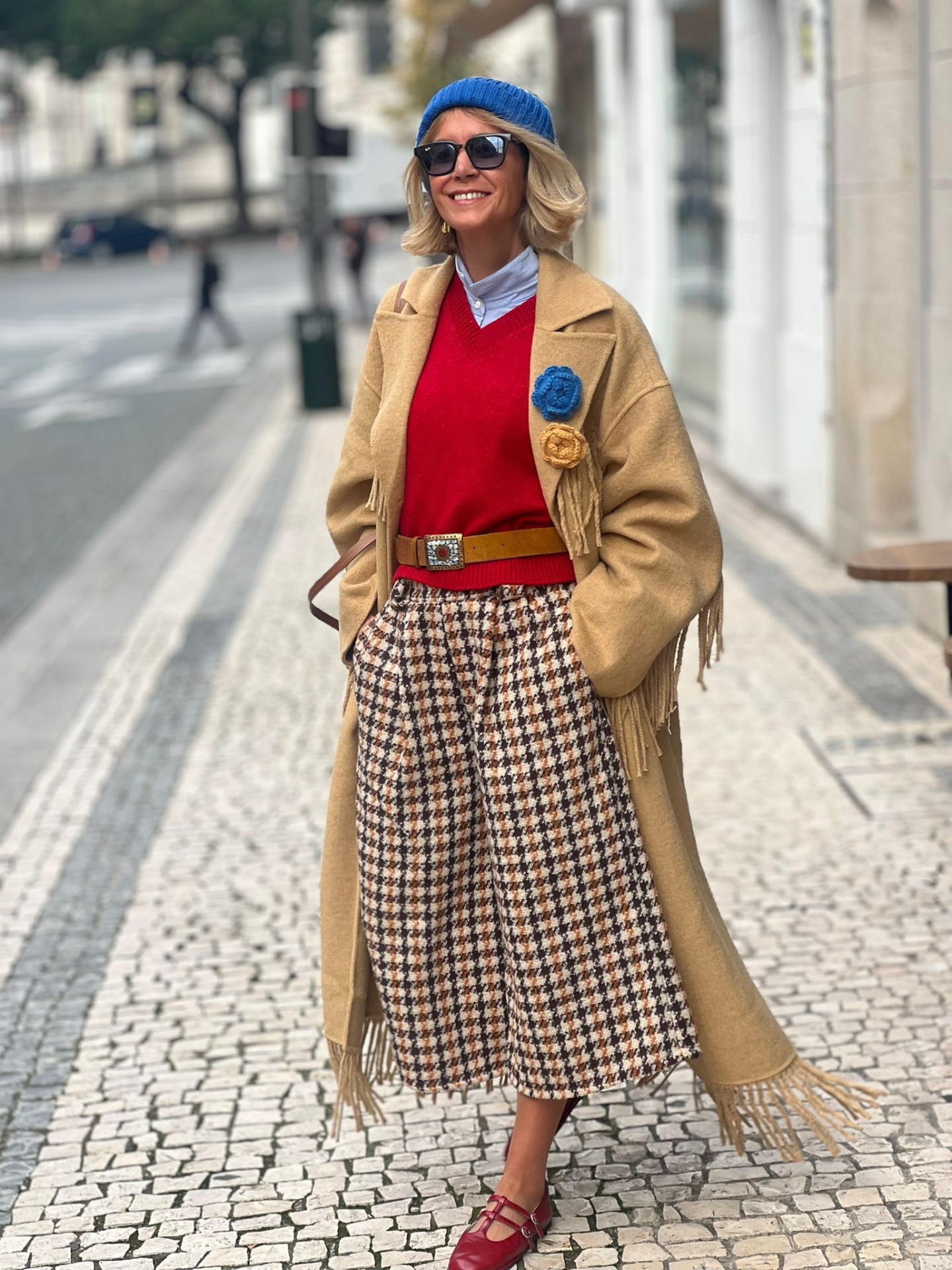 Mulher com casaco bege, camisola vermelha, saia pied-de-poule, chapéu azul, óculos escuros e sapatos vermelhos numa calçada urbana