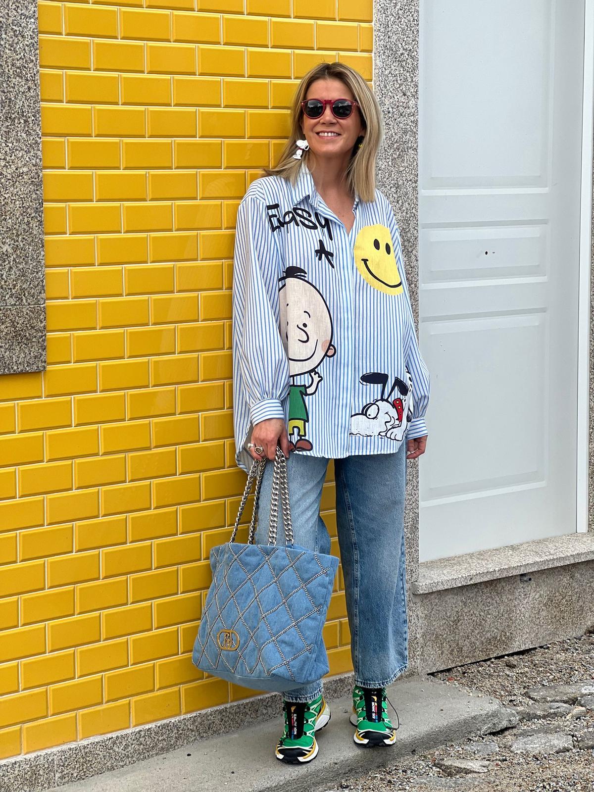 Pessoa vestindo camisa estampada, jeans, sapatilhas, óculos de sol e bolsa azul, em frente a parede amarela.