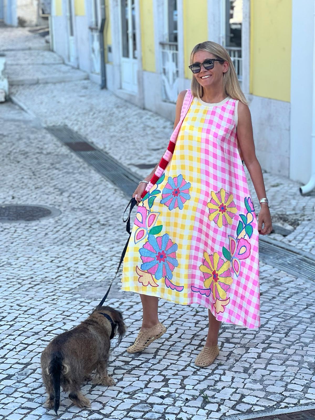 Mulher de vestido longo xadrez amarelo e rosa com flores coloridas, óculos escuros, andando com cão numa rua de calçada portuguesa.