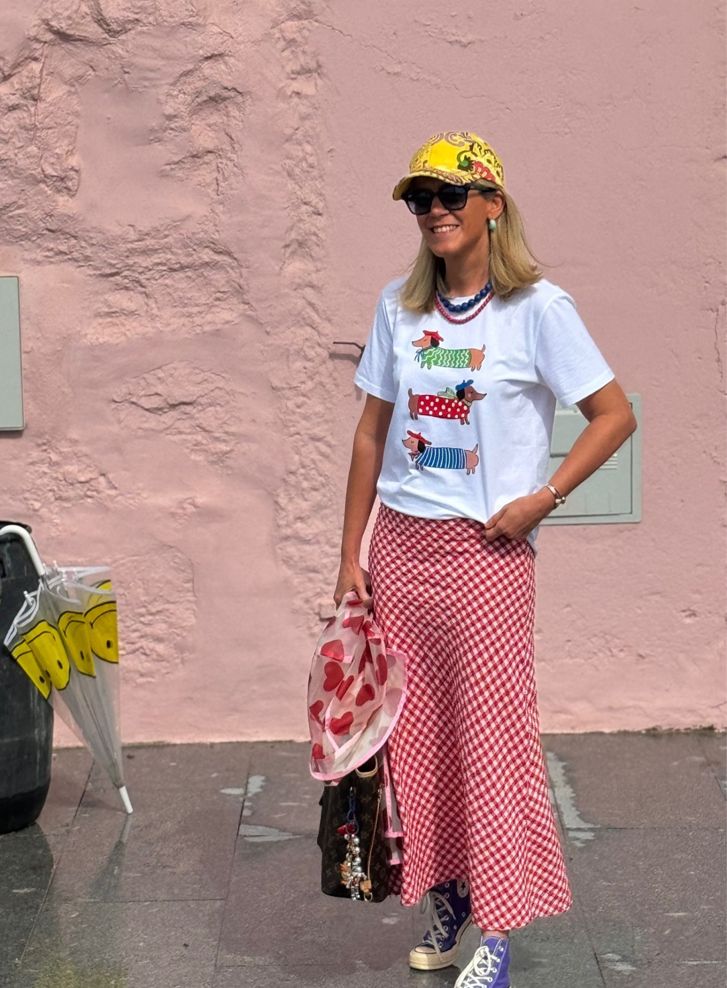 Mulher vestindo t-shirt branca e saia xadrez vermelha e branca, com boné amarelo e óculos escuros numa rua com parede rosa