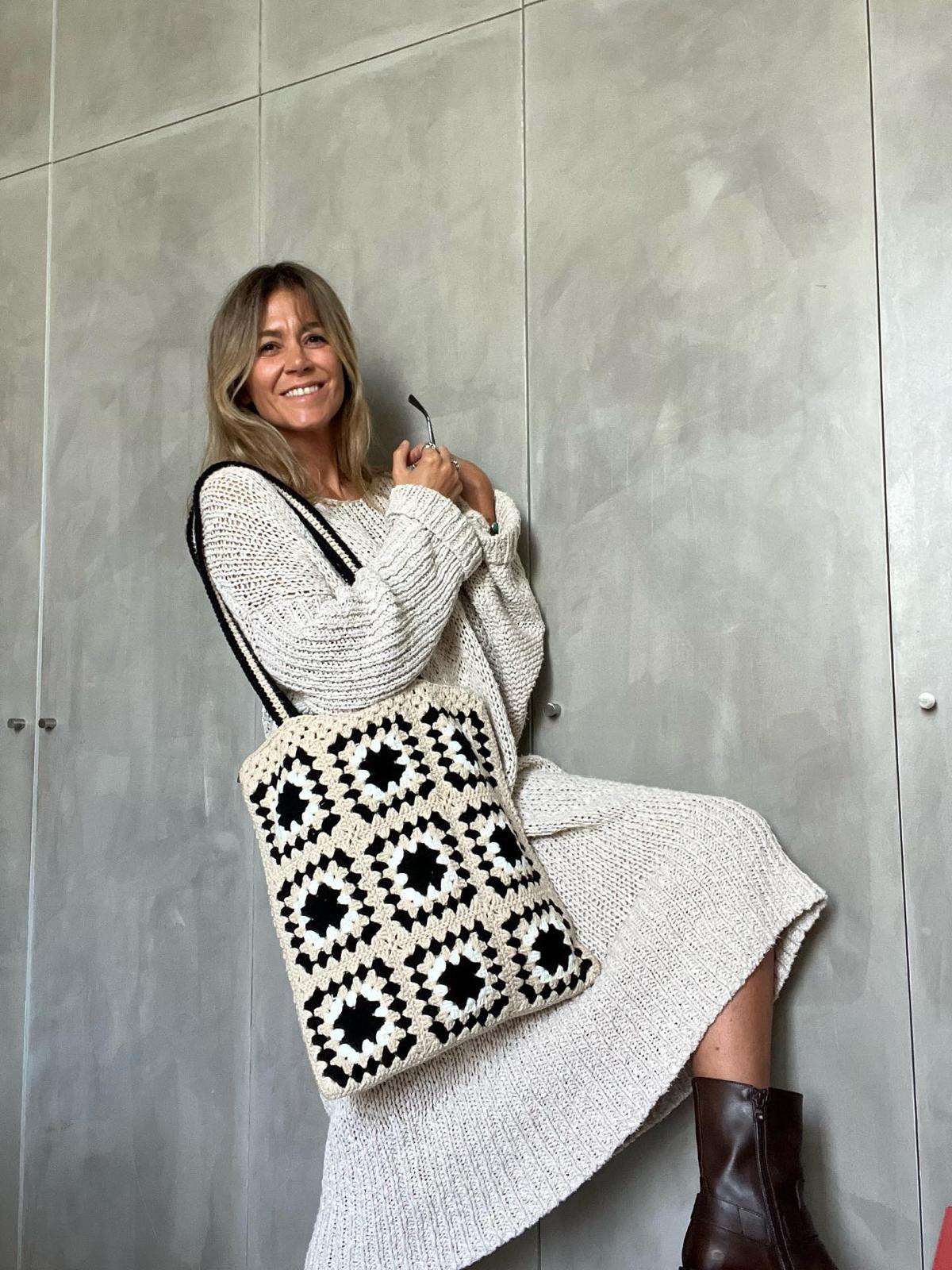 Mala de tricot bege com padrão de quadrados pretos junto a mulher com vestido branco e botas castanhas