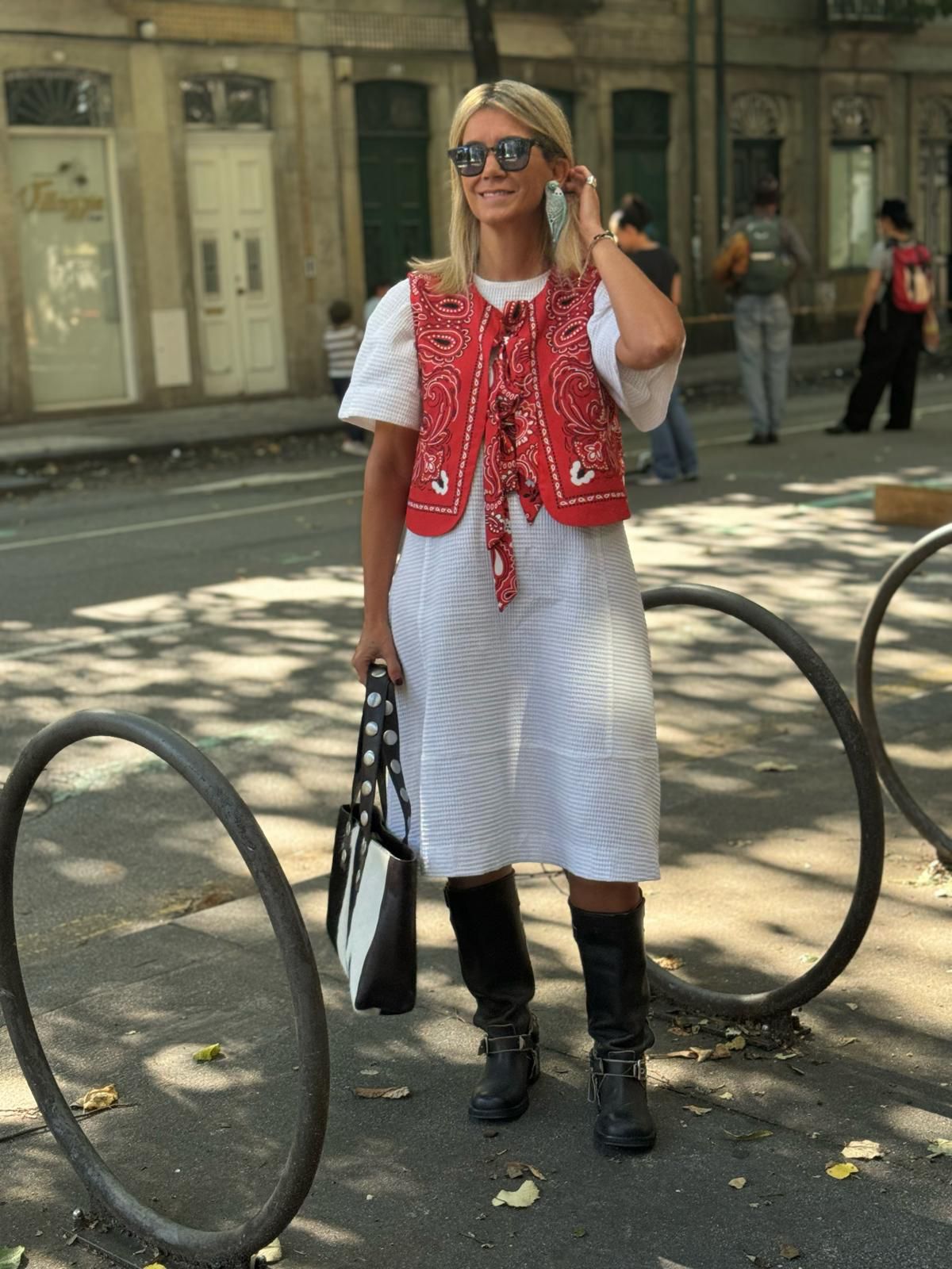 Mulher com vestido branco, colete vermelho estampado, botas pretas e óculos escuros numa rua urbana.