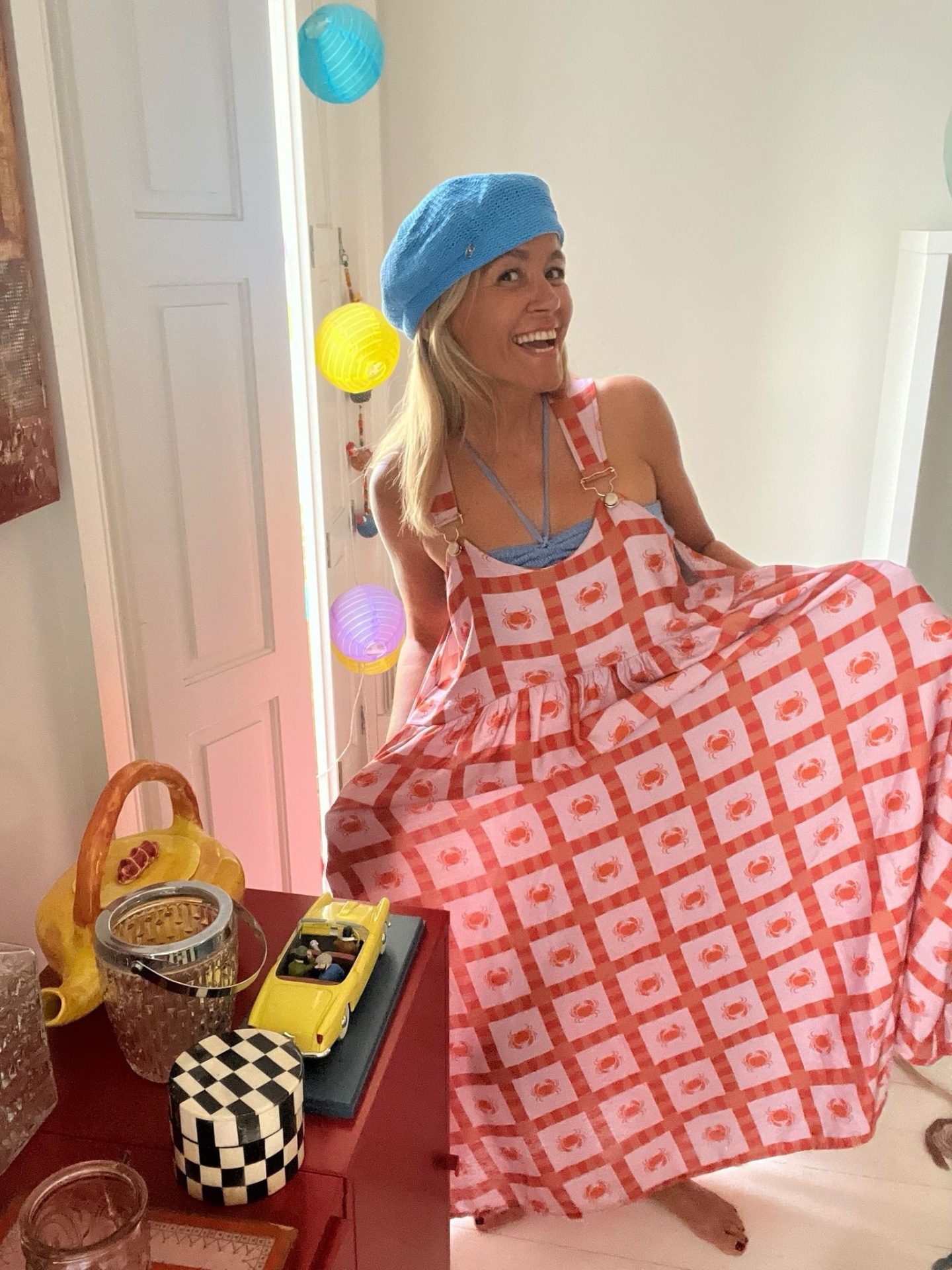 Vestido longo rosa com padrão quadriculado e flores vermelhas, boina azul, decoração interior colorida.