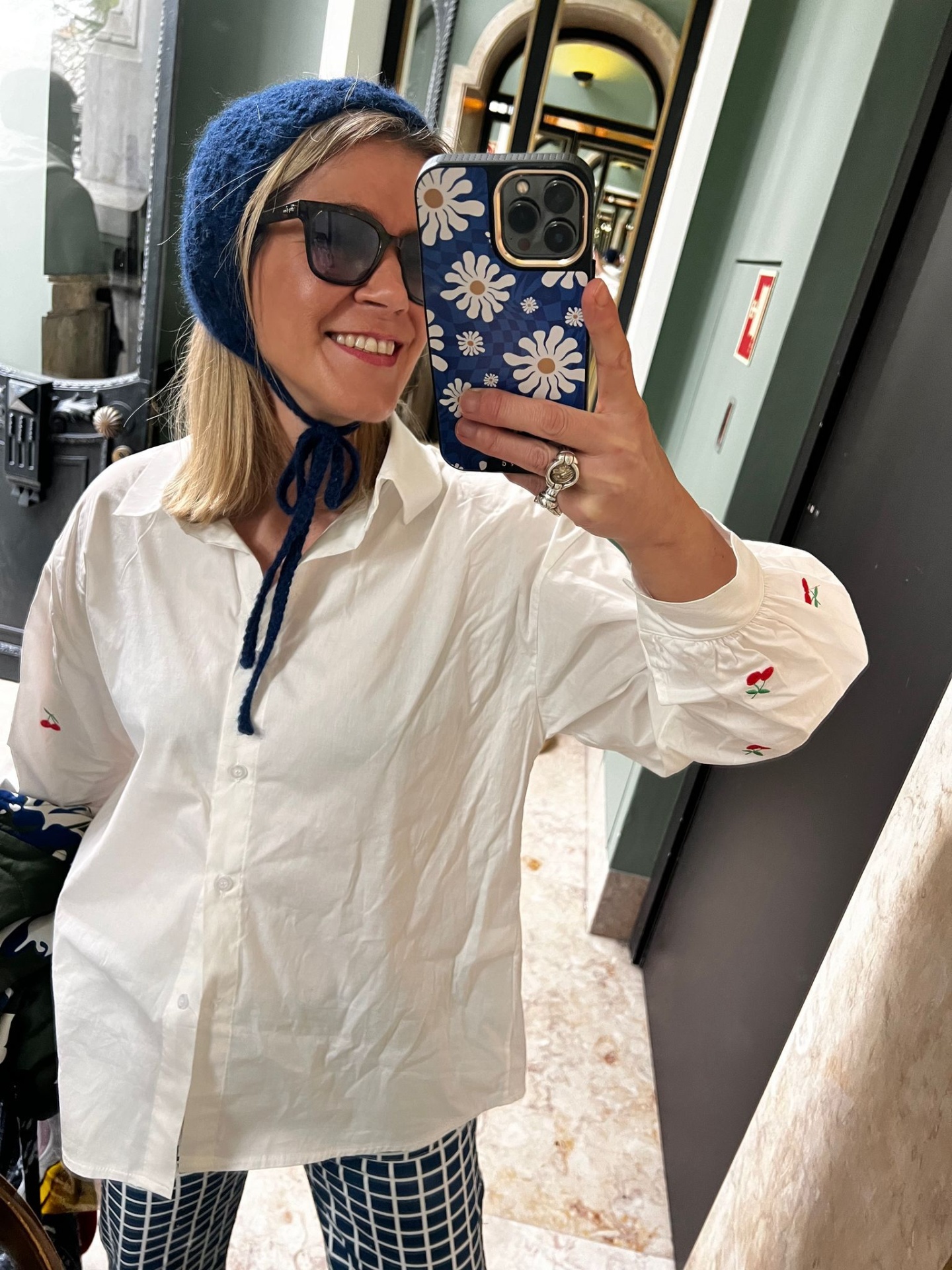 Mulher com camisa branca bordada, gorro azul, óculos escuros e capa de telemóvel azul com flores.