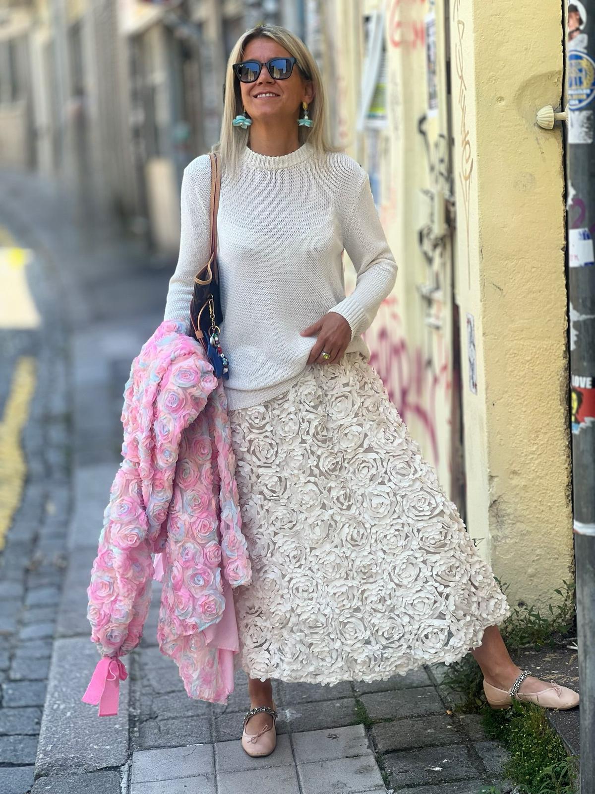 Mulher com camisola branca e saia com padrão de rosas, segurando casaco rosa com flores, numa rua urbana