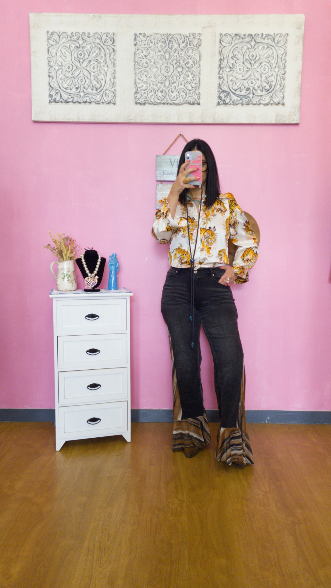 Pessoa vestindo blusa com padrão de tigres e calças pretas com barra animal print, em frente a parede cor de rosa com quadro e móvel branco