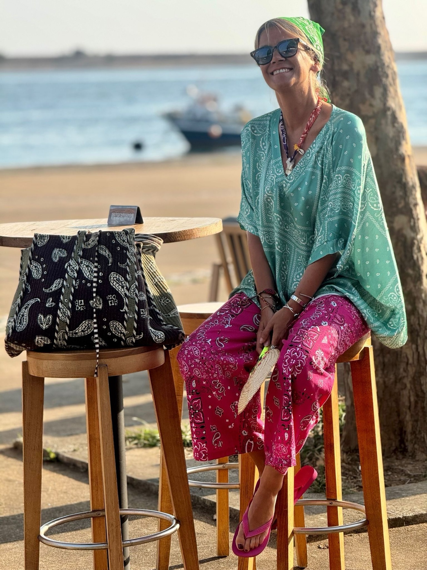 Mulher com roupa colorida numa esplanada à beira-mar com mala preta e barco ao fundo.