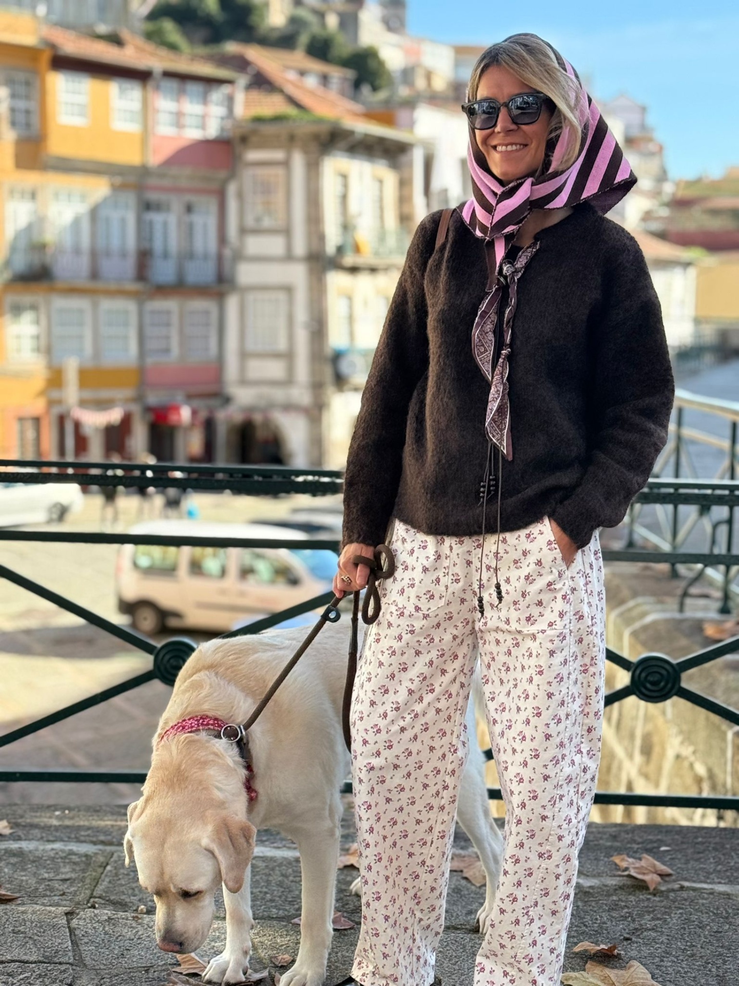 Mulher com casaco castanho e calças brancas florais a passear cão Labrador creme numa rua urbana