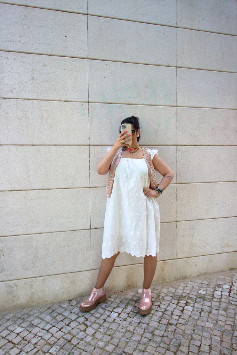Mulher de vestido branco, colete bege, botas cor de rosa, posando contra parede branca
