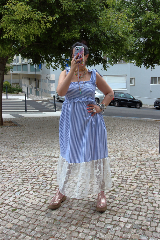 Mulher com vestido azul às riscas e renda branca abaixo, sandálias rosa dourado, tirando selfie num passeio de pedra com árvores e carros ao fundo