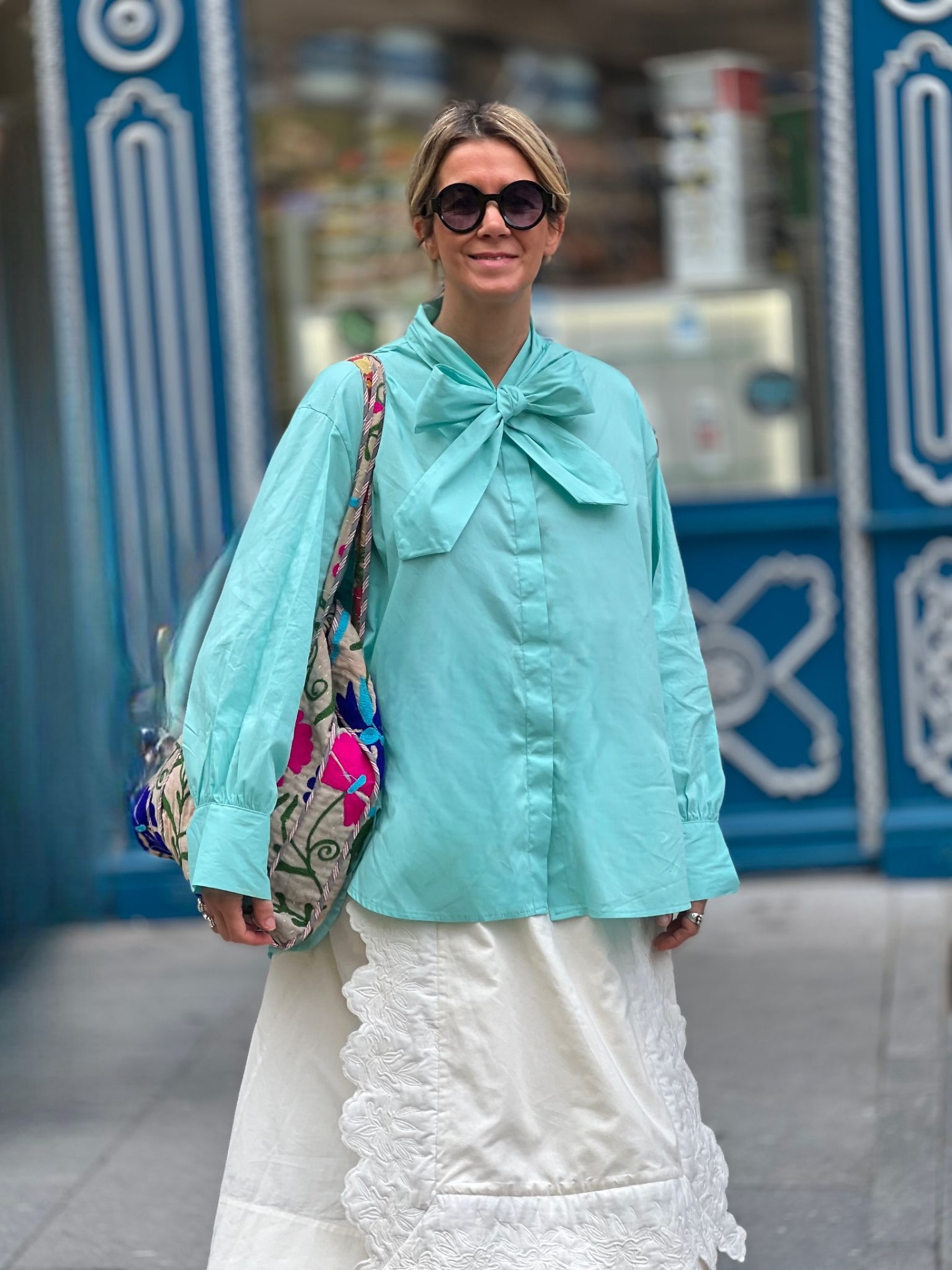 Mulher com camisa azul clara, saia branca rendada, mochila floral e óculos de sol pretos na rua