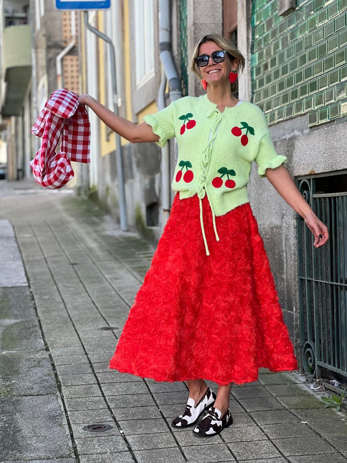 Mulher com casaco verde claro com cerejas, saia vermelha texturizada e sapatos preto e branco numa rua de calçada
