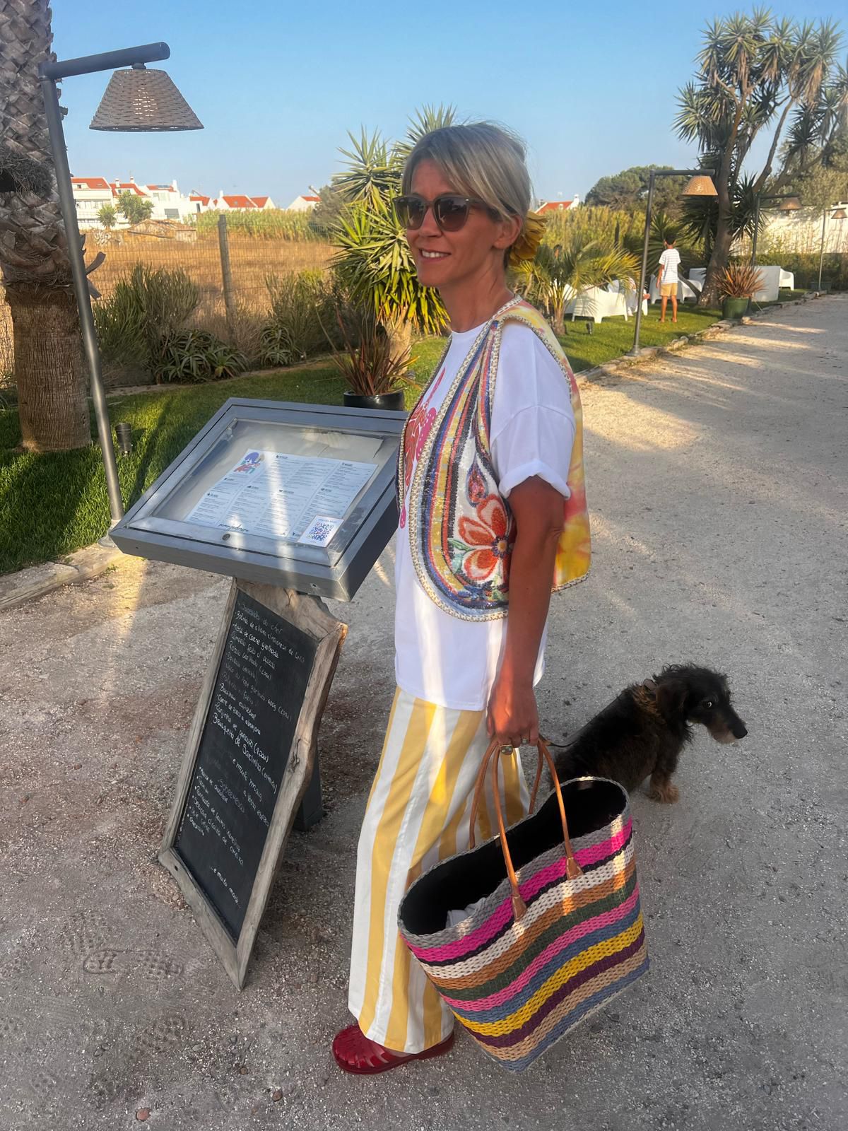 Mulher com roupa colorida e sacola de palha segura ao lado de menu metálico em ambiente exterior com cão próximo