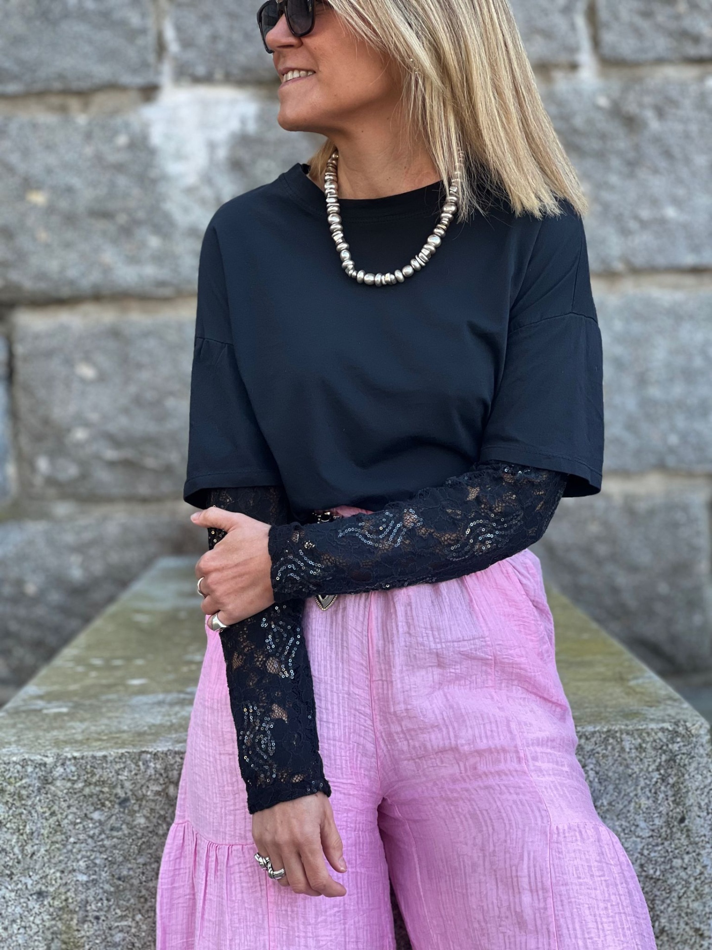 Mulher com camiseta preta, camisa rendada preta, calças rosas largas, colar de contas prateadas, óculos escuros, encostada em parede de pedra.