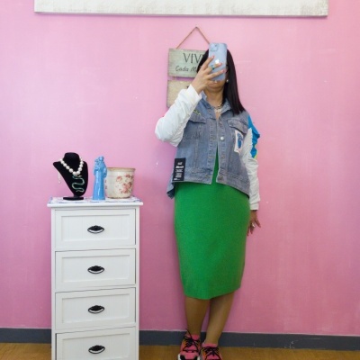 Pessoa tirando selfie com vestido verde e colete jeans em frente a parede rosa com decoração e móvel branco com bijuterias.
