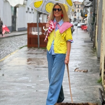 Mulher com guarda-chuva transparente com carinhas sorridentes, colete amarelo e calças azuis em rua molhada