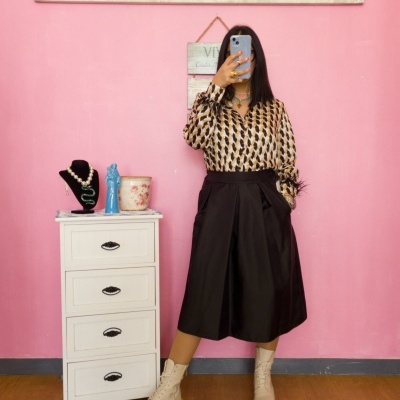 Mulher vestida com camisa estampada e saia preta, botins bege, em frente a parede cor de rosa com móvel branco e decoração