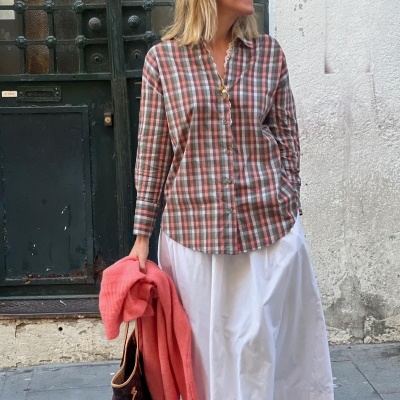Pessoa com camisa xadrez castanha e rosa e saia branca, segurando casaco coral e mala castanha