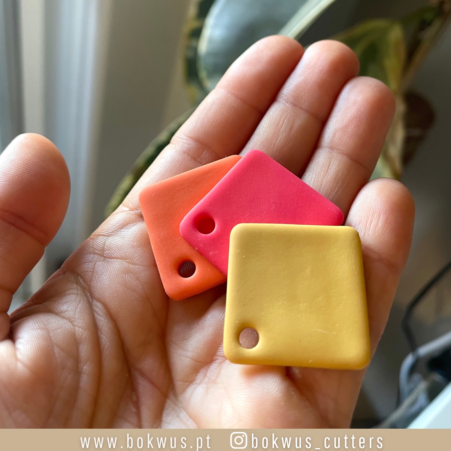 Etiquetas quadradas em silicone laranja, rosa e amarela numa mão com planta ao fundo