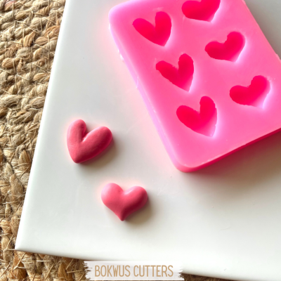 Forma de silicone rosa com cavidades de coração, dois corações moldados em pasta rosa, sobre superfície branca e esteira natural com texto BOKWUS CUTTERS