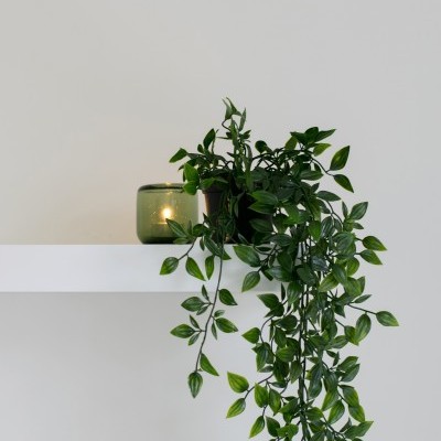 Planta artificial verde pendurada numa prateleira branca, com copo verde e vela acesa ao lado