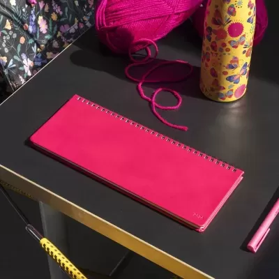 Caderno espiral rosa, bola de lã, garrafa decorada, caneta sobre mesa preta com bordas douradas