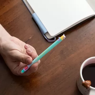Lápis azul decorativo sendo segurado por uma mão sobre mesa de madeira, com caderno e chávena ao fundo.