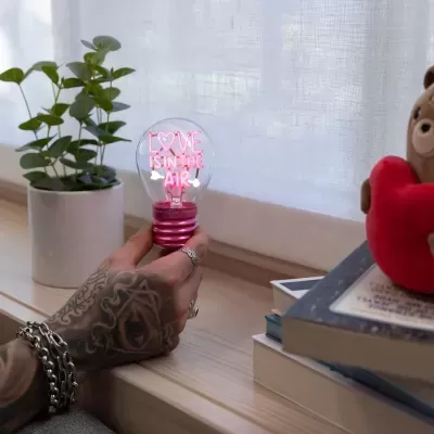 Lâmpada decorativa com texto LOVE IS IN THE AIR segurada por mão tatuada