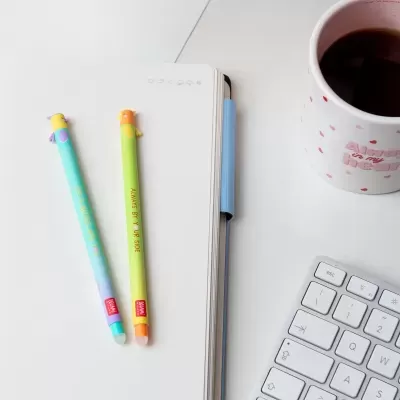 Dois lápis coloridos com passarinho, caderno aberto, caneca rosa e teclado branco em fundo branco
