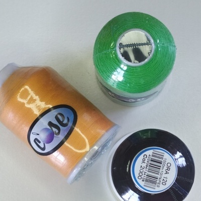 Três rolos de linha de costura nas cores verde, laranja e preta com etiquetas visíveis