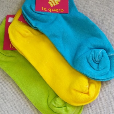 Três pares de meias coloridas azul, amarelo e verde com etiquetas rosas sobre tecido cinza.