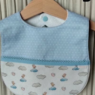 Babete azul claro com padrão de pontos brancos e padrão de nuvens e passarinhos, pendurado em cabide de madeira.
