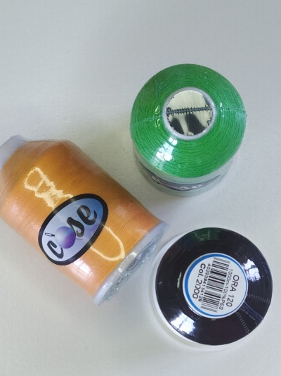 Três rolos de linha de costura nas cores verde, laranja e preta com etiquetas visíveis
