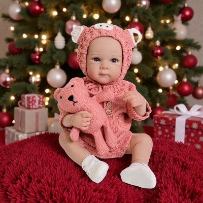 Boneca realista com roupa rosa e urso de peluche sentada em almofada vermelha com árvore de Natal ao fundo
