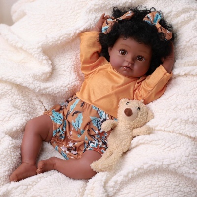 Boneca realista de criança vestida com camisola laranja e calções com padrão azul e castanho sobre manta branca felpuda com urso de peluche.