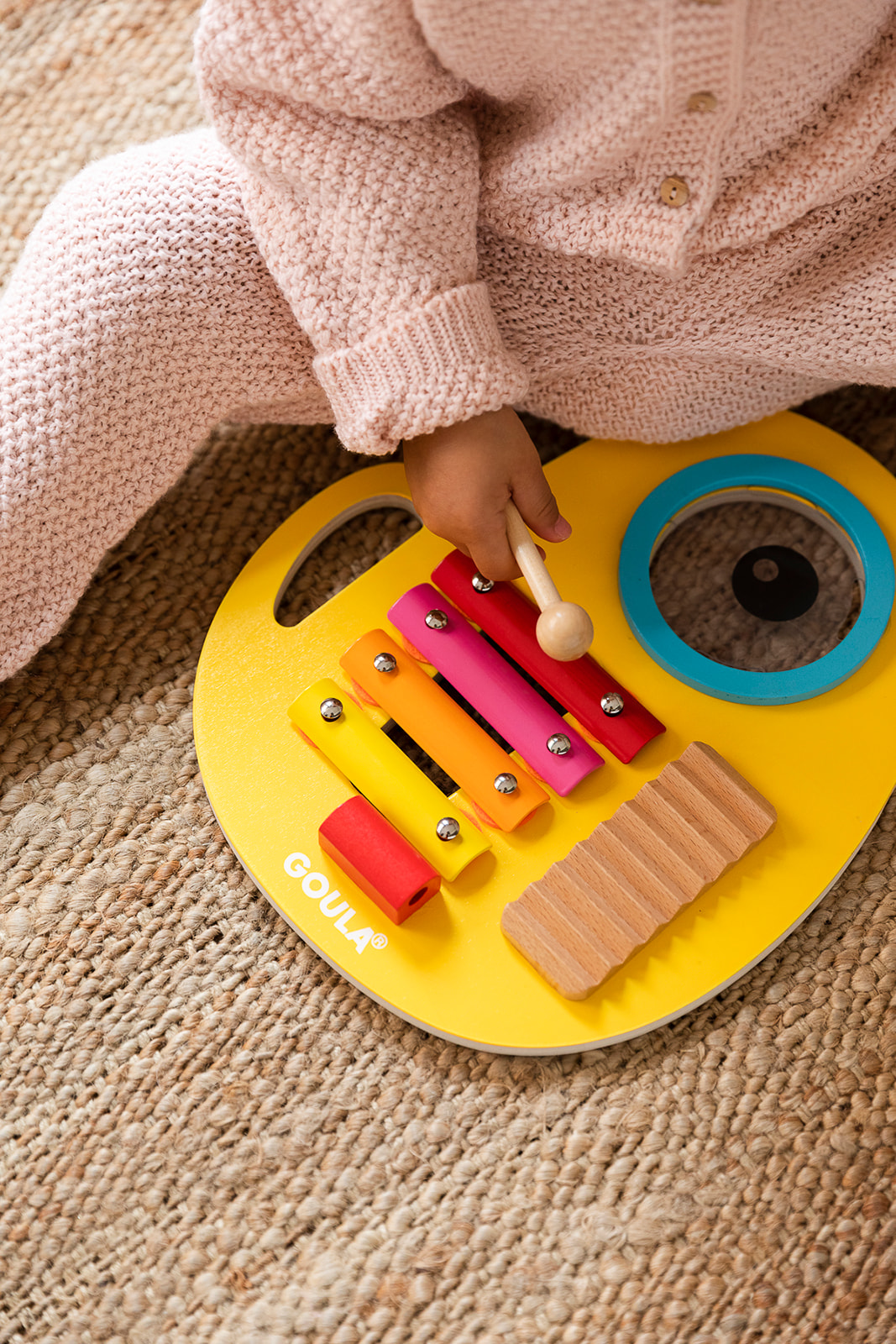 TIKI MUSICAL - BRINQUEDO MUSICAL 3 EM 1, GOULA Percussão infantil de madeira e metal amarela com barras coloridas e criança com roupa rosa sentada em tapete de fibras.