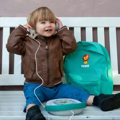 Criança com casaco castanho e auscultadores sentada em banco branco com mochila verde TIMIO ao lado
