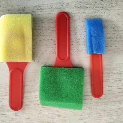 Três pincéis de esponja com cabos vermelhos e esponjas nas cores amarelo, verde e azul sobre superfície de madeira clara