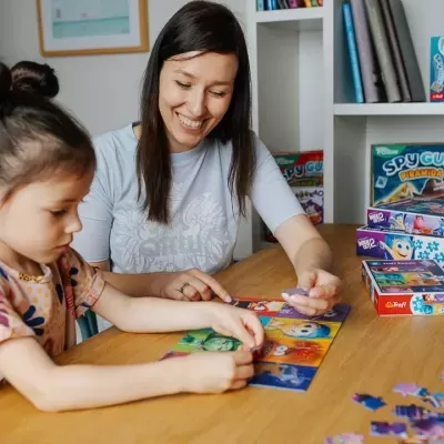Criança e mulher montando um quebra-cabeças infantil de papel com personagens, com caixas de jogos ao fundo