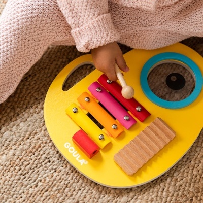 Percussão infantil de madeira e metal amarela com barras coloridas e criança com roupa rosa sentada em tapete de fibras.