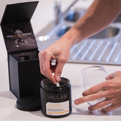 Frasco preto de suplemento PILLAR Ultra Immune C com comprimido a ser retirado e copo transparente perto de pia