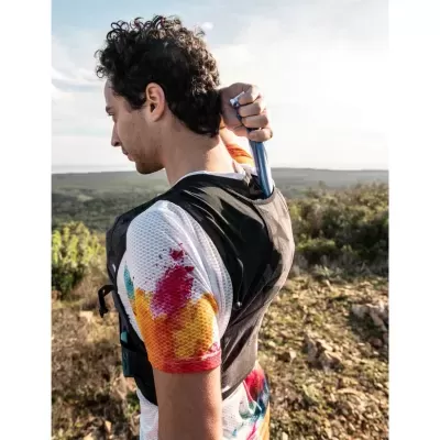 Homem veste colete preto e t-shirt colorida, segura cilindro metálico atrás das costas em ambiente natural