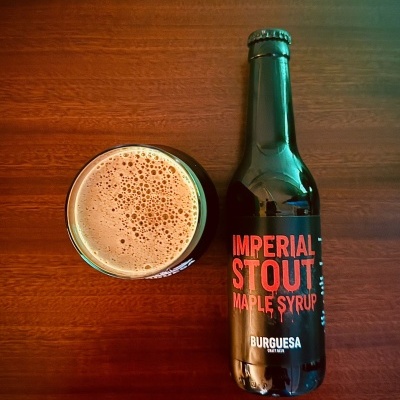 Garrafa de cerveja artesanal escura com rótulo Imperial Stout Maple Syrup Burguesa e copo com cerveja foam em mesa de madeira.