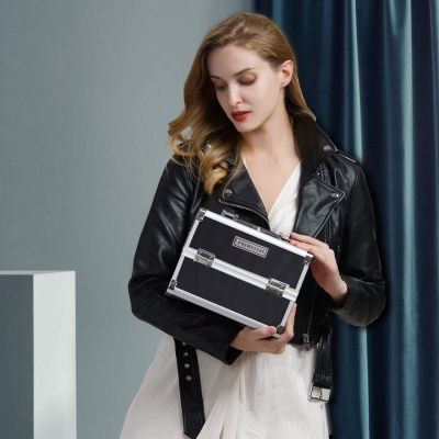 Mulher loira veste casaco de couro preto e vestido branco segurando mala preta rígida com armações metálicas