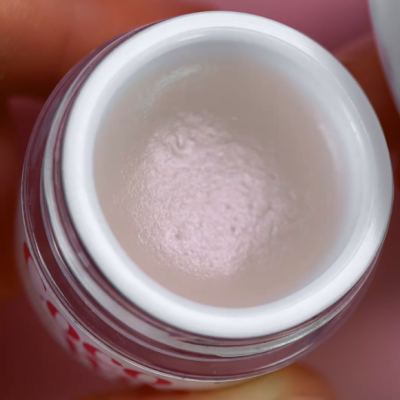 Frasco aberto com creme ou pomada creme, segurado por dedos, fundo rosa desfocado