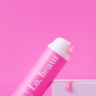 Frasco rosa de esfoliante labial Lo. beaut com creme e caixa branca num fundo rosa