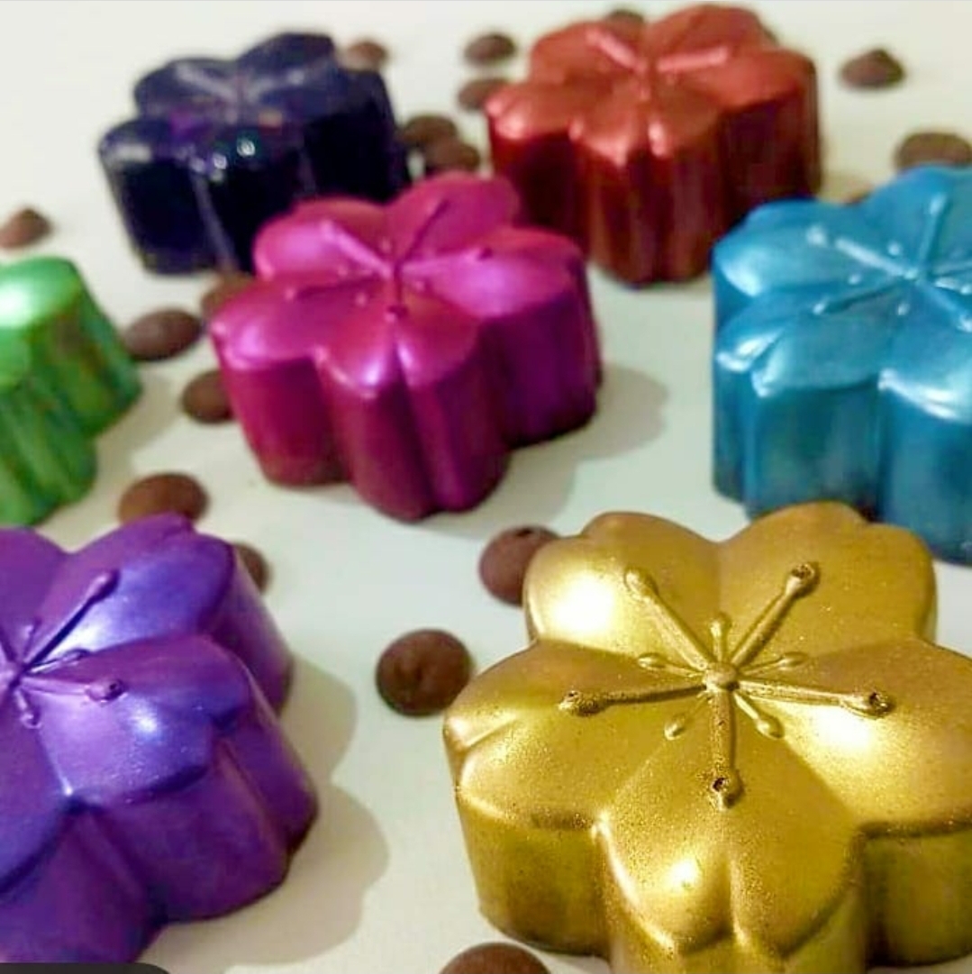 Oito chocolates em forma de flor coloridos com acabamento metálico brilhante sobre fundo branco com grãos de café.
