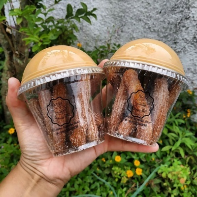 Dois copos transparentes de plástico com churros e tampa amarela em mão, fundo natural verde com flores amarelas