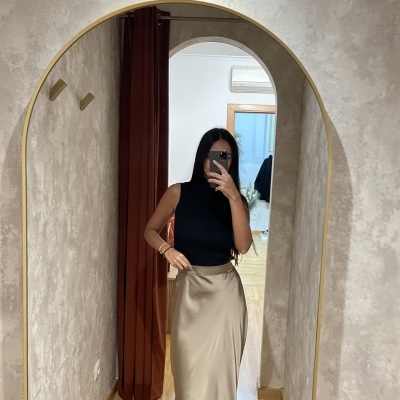 Mulher com saia nude de cetim e top preto refletida em espelho de arco com moldura dourada