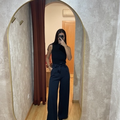 Mulher a tirar uma selfie num espelho oval, vestindo colete preto e calças pretas largas